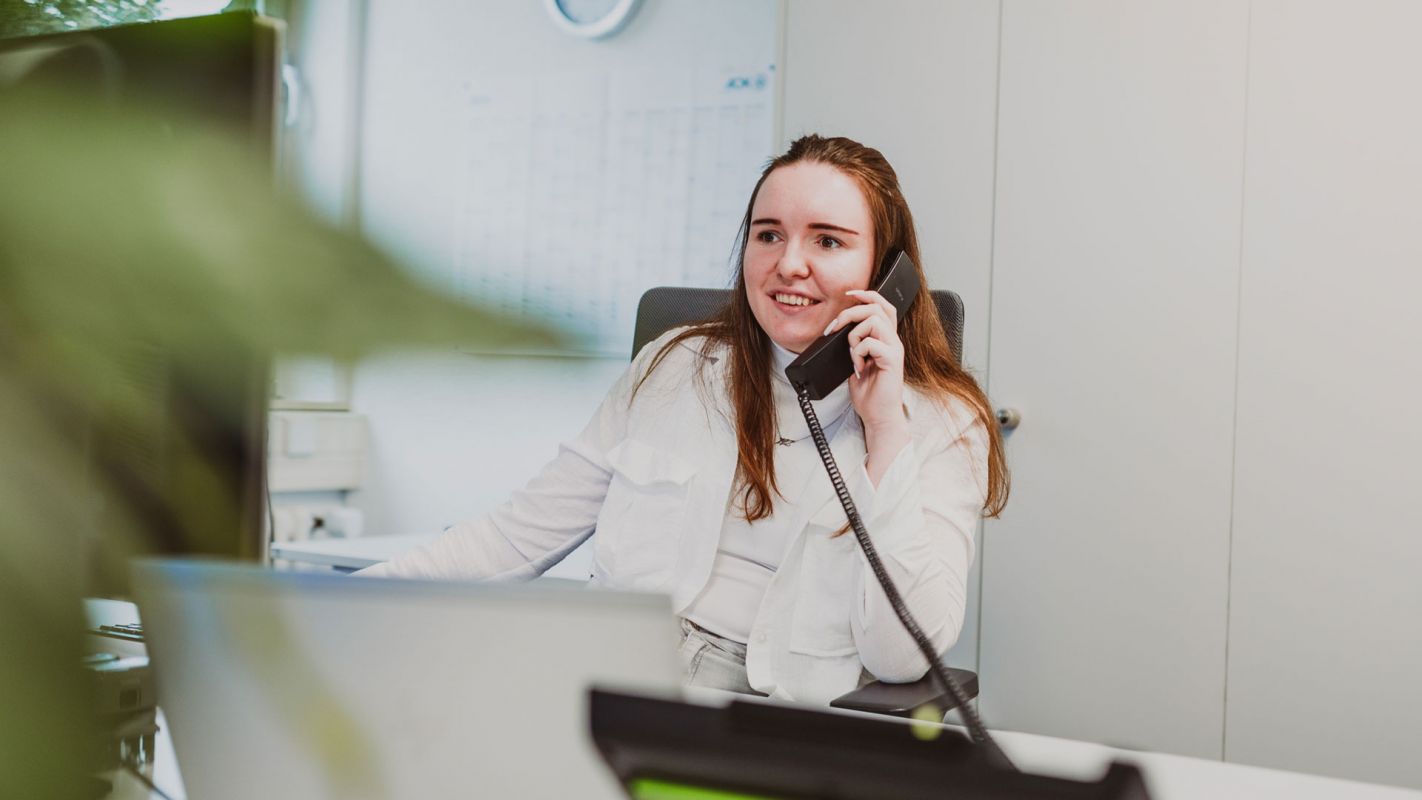 Kontaktaufnahme mit Mitarbeiterin von Weber. Junge Ansprechpartnerin von Weber spricht am Telefon vor einem Rechner. Im Vordergrund eine unscharfe Pflanze, im Hintergrund eine helle Bürougebung.